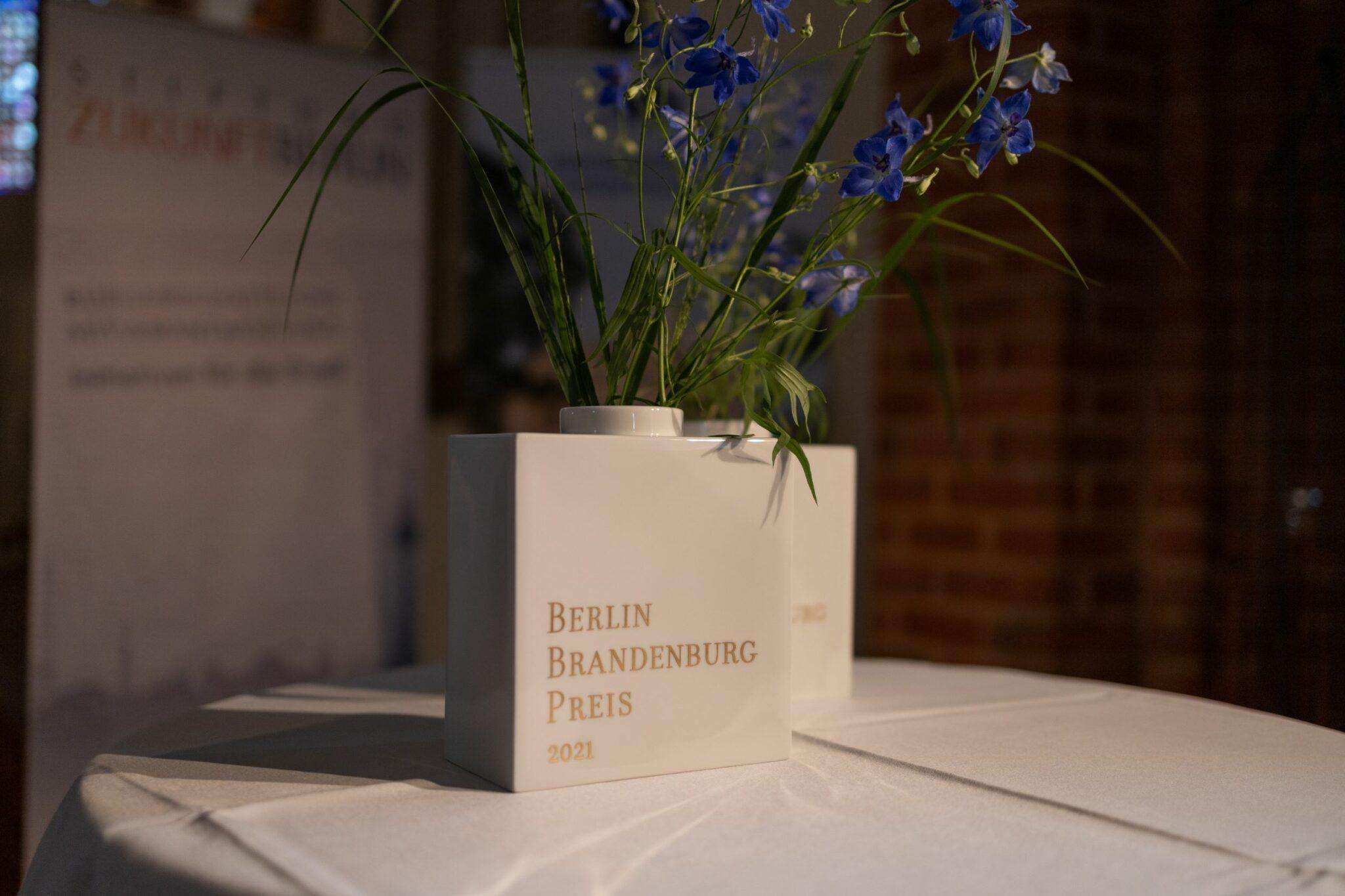 Zwei weiße Porzellanvasen mit dem goldenen Schriftzug BERLIN BRANDENBURG PREIS und Blumensträußen.