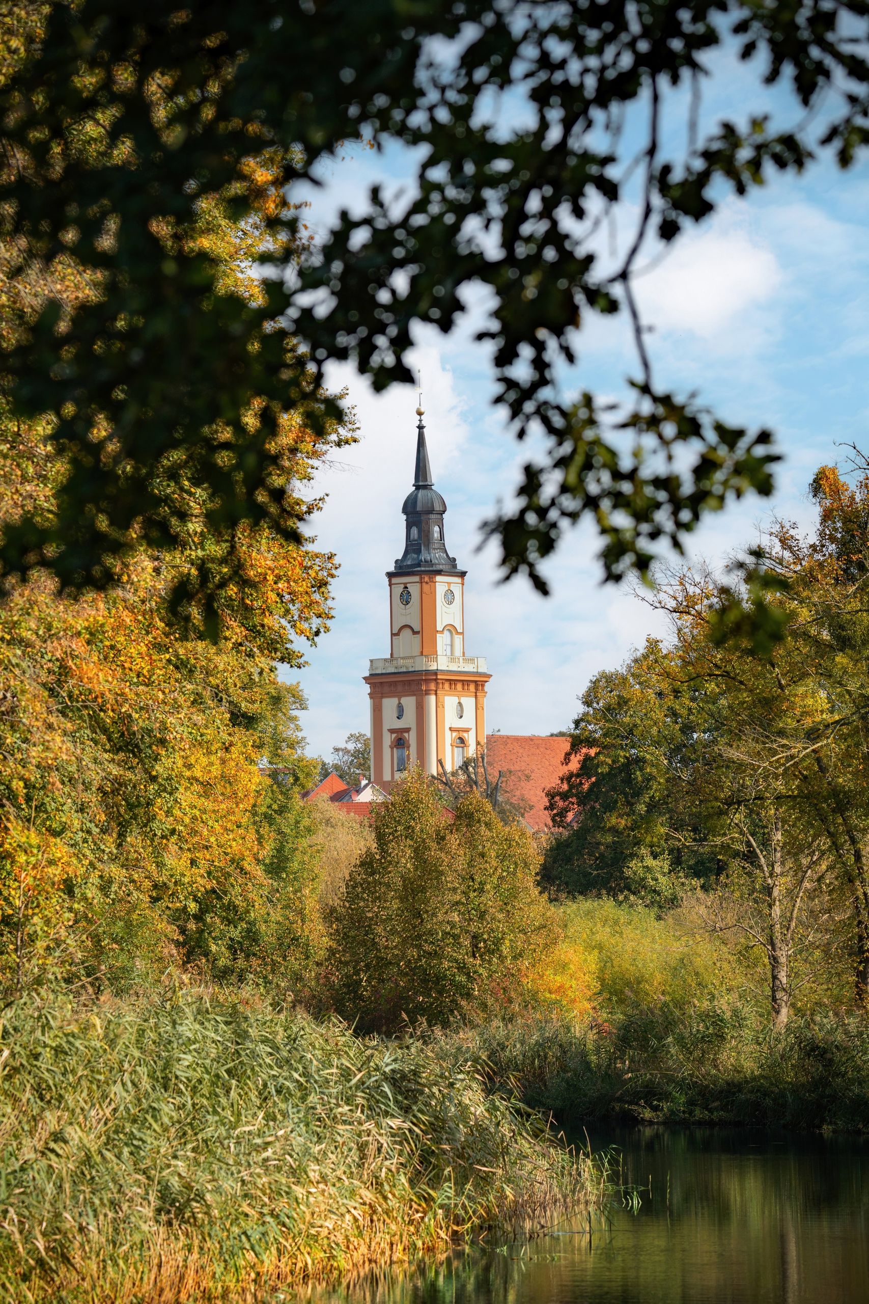 A scenic view of a Gothic clock tower surrounded by autumn trees beside a canal.
